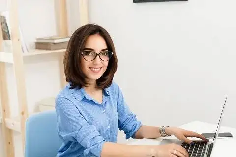 pretty brunette girl is sitting typing laptop table office she is smiling friendly camera she wears blue shirt black glasses